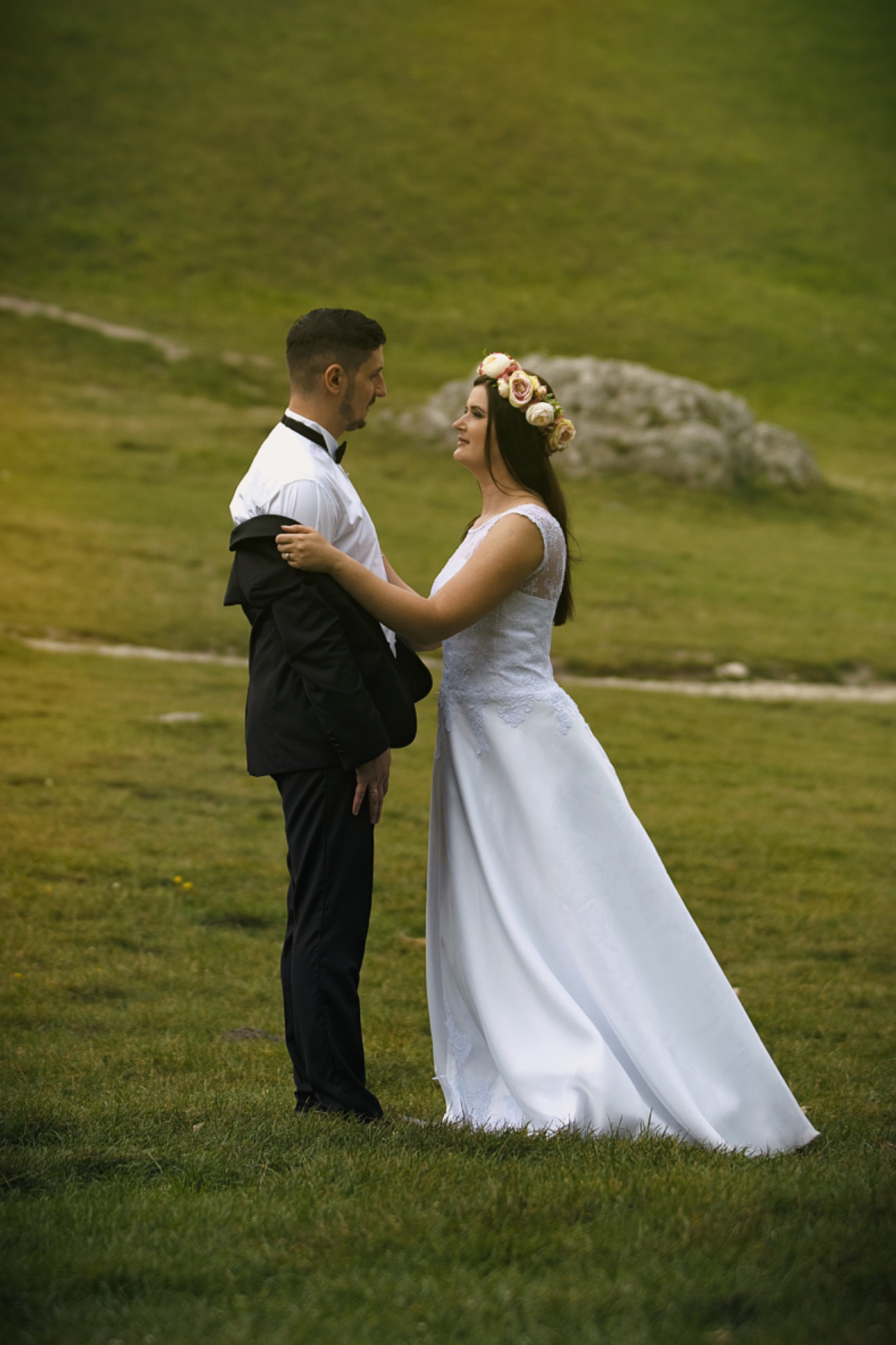 Para młoda w strojach ślubnych na tle zielonego wzgórza, panna młoda z wiankiem na głowie zakłada marynarkę na pana młodego.
