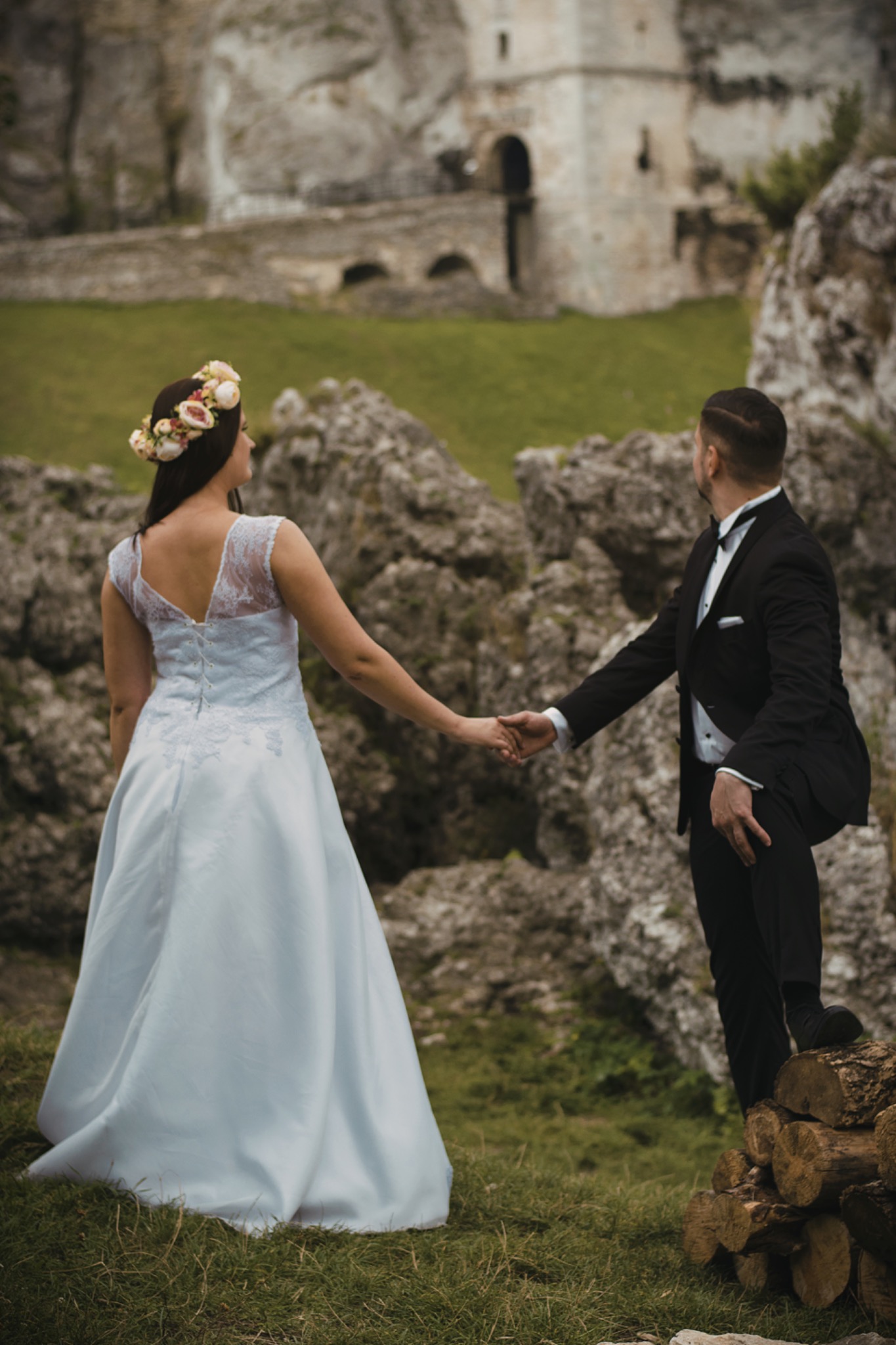 Para młoda trzymająca się za ręce, odwrócona plecami do widza, na tle zamku w ruinie i skalistego krajobrazu; panna młoda w sukni z koronkowymi plecami i wiankiem na głowie, pan młody w smokingu...