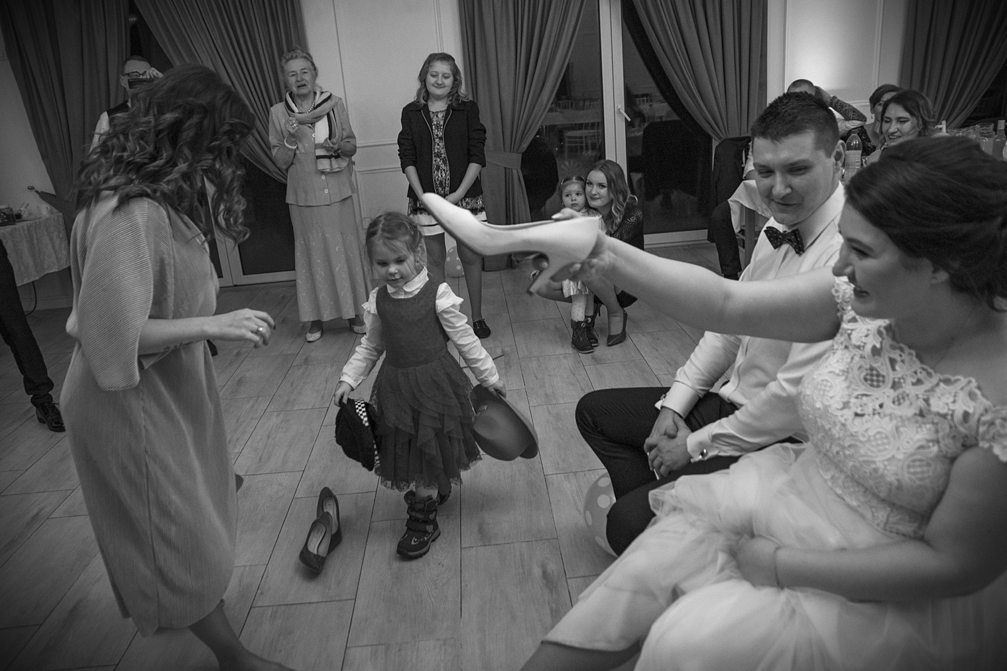 Czarno-białe zdjęcie z wesela: panna młoda siedzi, trzyma szpilkę w ręku, obok stoi mała dziewczynka w sukience, trzymająca kapelusz, na parkiecie leżą buty, w tle goście.