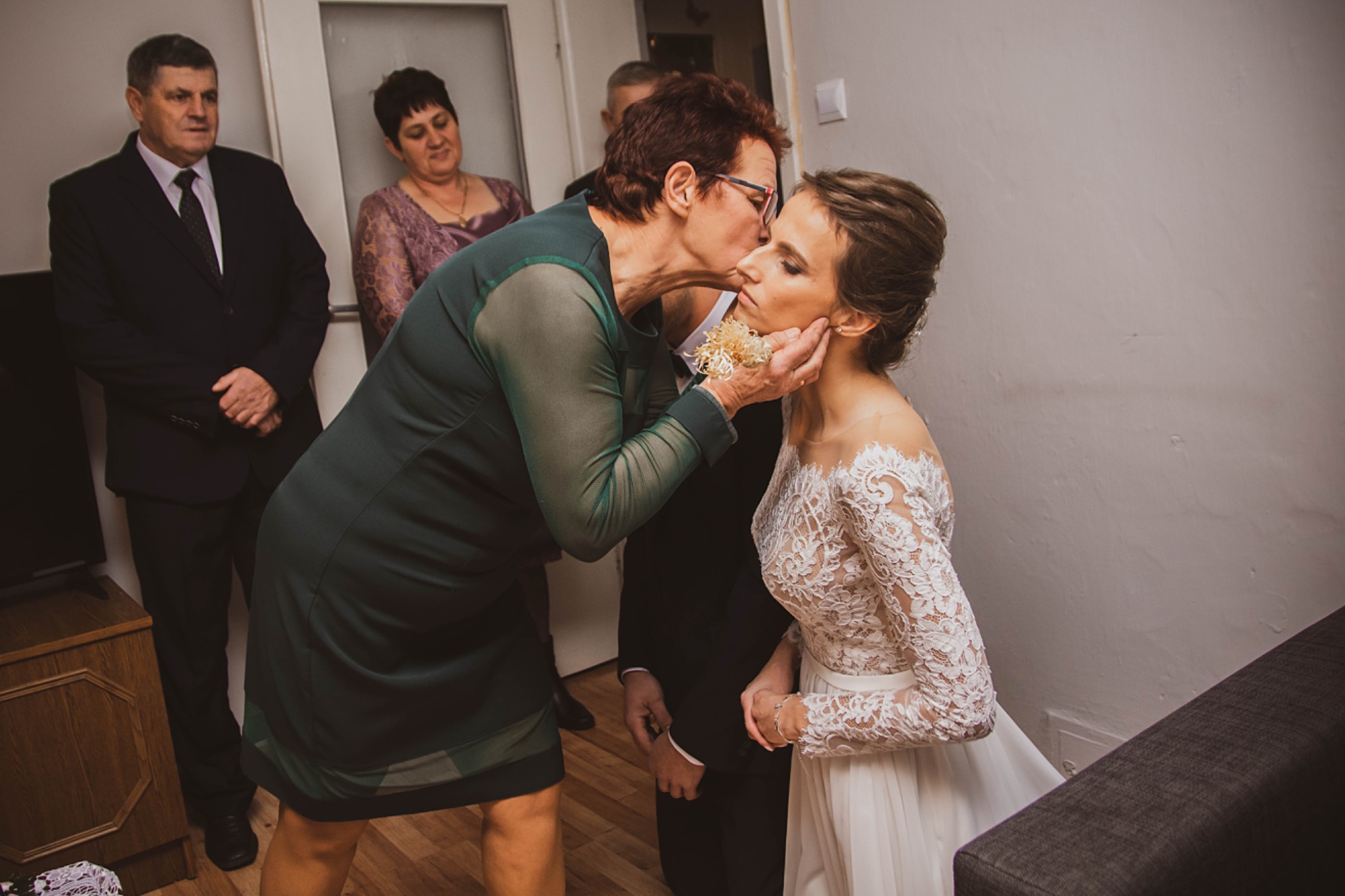 Wzruszający moment: kobieta w zielonej sukience całuje pannę młodą w koronkowej sukni, w tle obserwujący goście.