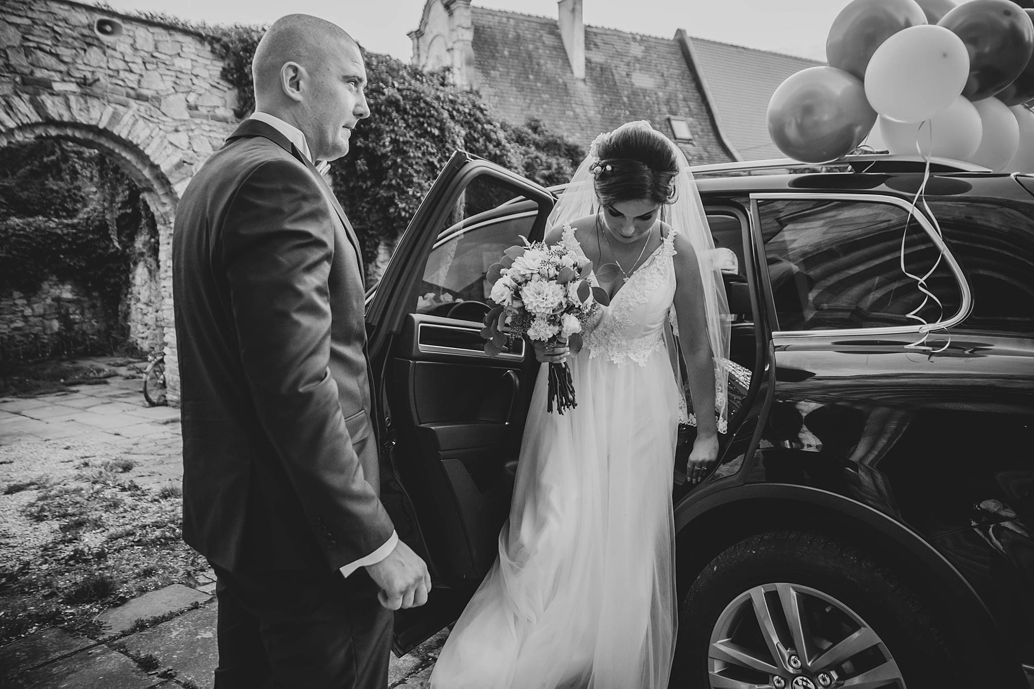 Czarno-białe zdjęcie panny młodej w sukni ślubnej wysiadającej z czarnego samochodu ozdobionego balonami, obok stoi pan młody w garniturze, w tle kamienny budynek.