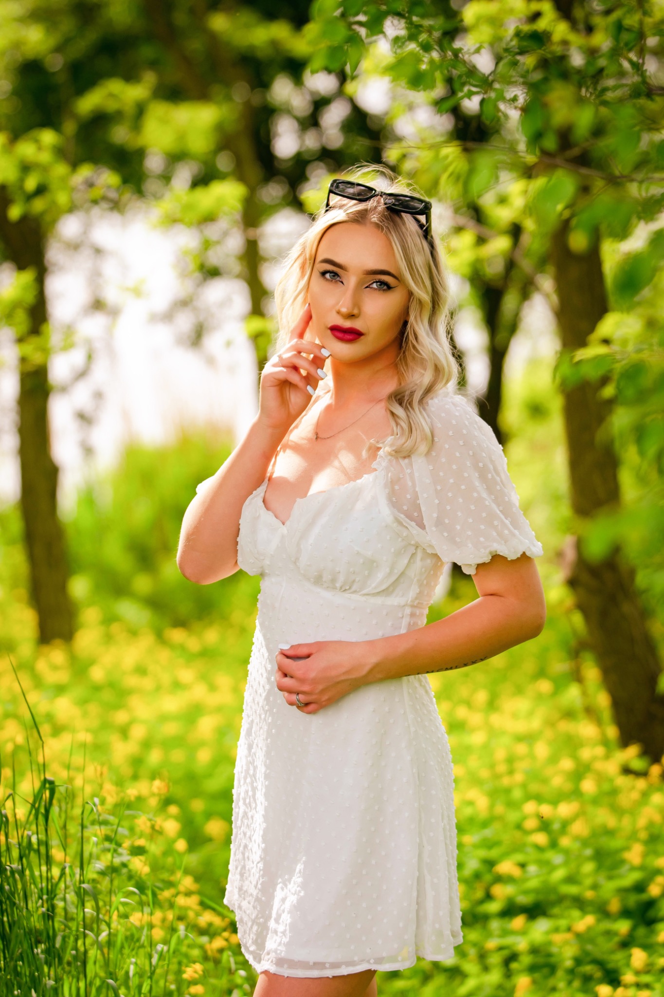 Portret kobiety w białej sukience na tle kwitnącej łąki z żółtymi kwiatami, okulary na głowie, delikatny uśmiech, naturalne światło.