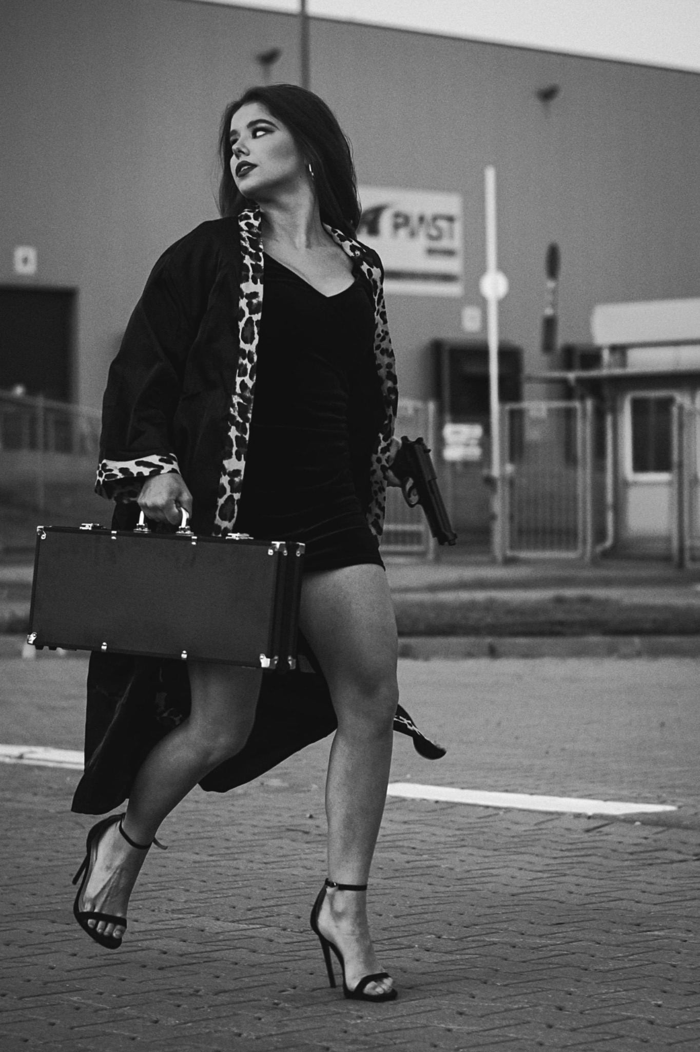 Czarno-białe zdjęcie kobiety w sukience i szlafroku w panterkę, biegnącej z walizką w jednej ręce i pistoletem w drugiej, na tle budynku.