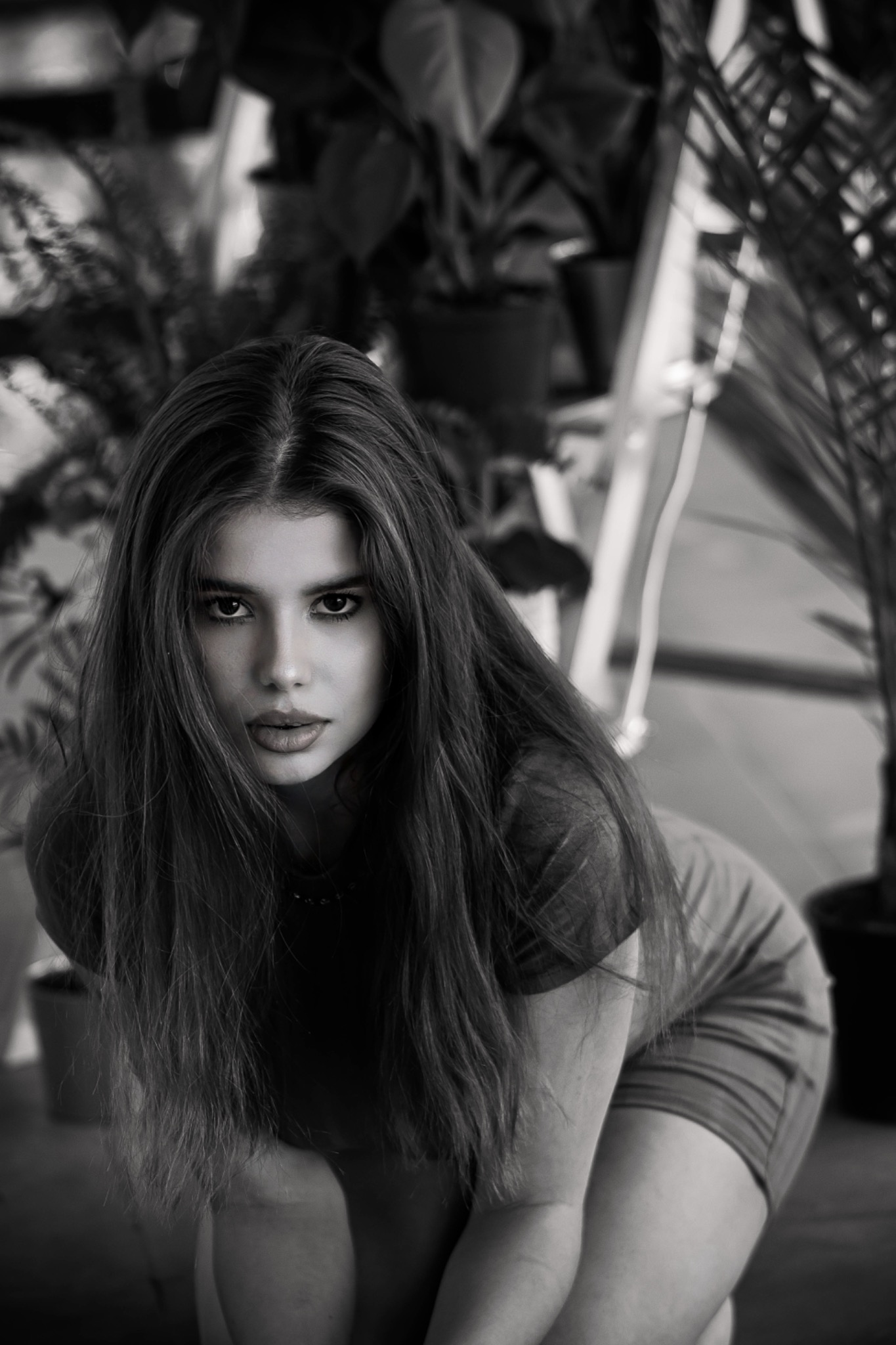 Portret młodej kobiety w czerni i bieli, pochylonej do przodu, z długimi włosami i intensywnym spojrzeniem, na tle roślin doniczkowych i drabiny.