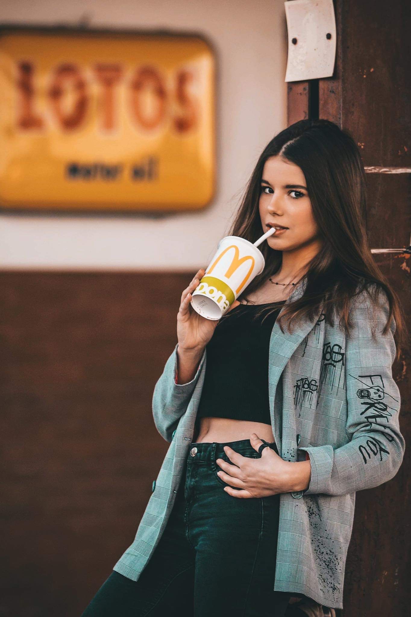 Młoda kobieta pije napój ze słomką z kubka McDonald's, ubrana w czarny top i szarą marynarkę w kratę z graffiti, opierając się o rdzewiejącą metalową konstrukcję na tle żółtego szyldu.