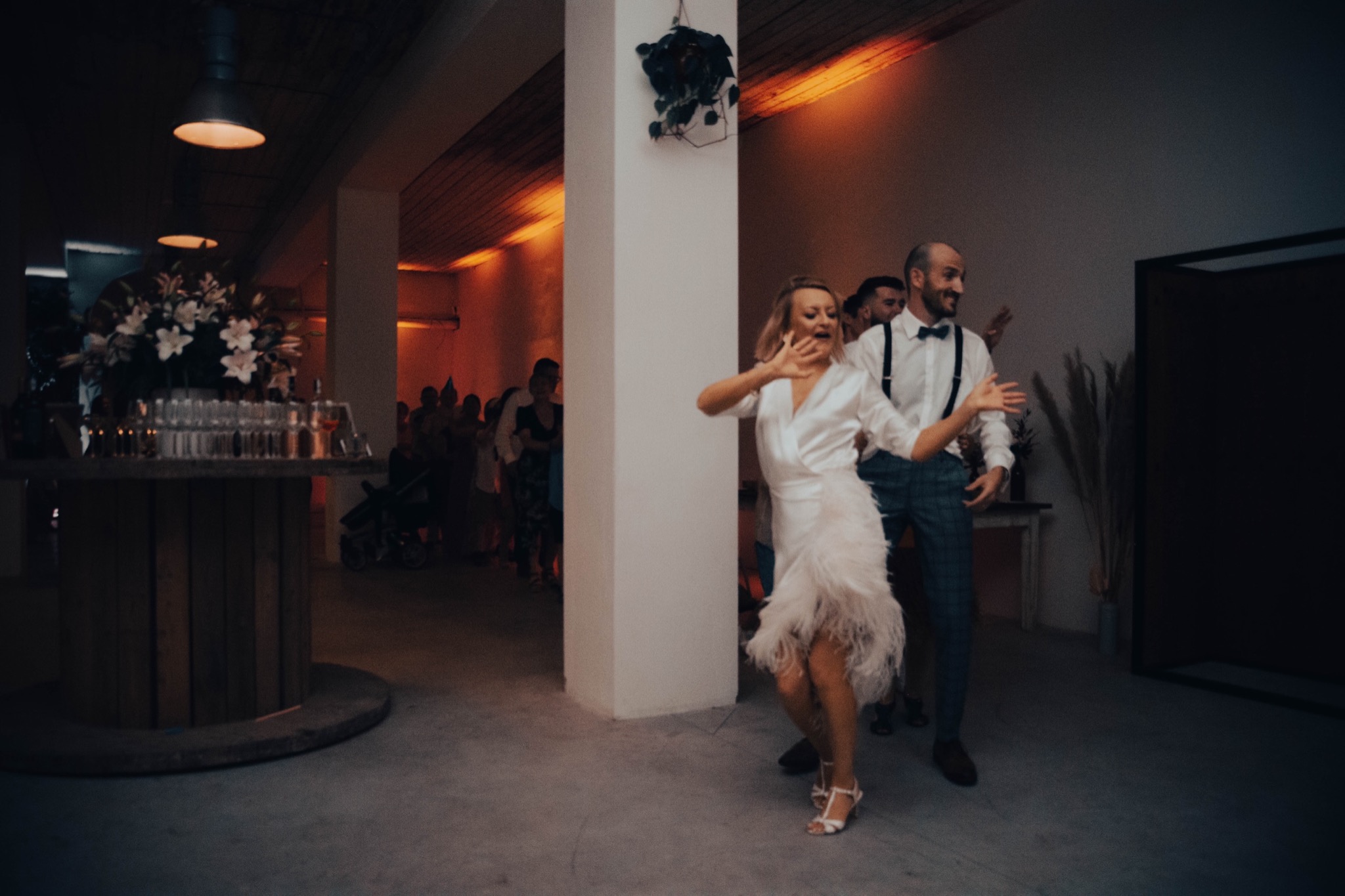 Para młoda tańczy podczas przyjęcia weselnego w przestronnym wnętrzu z betonową podłogą, kolumną i dekoracjami kwiatowymi, oświetlonym ciepłym światłem lamp.