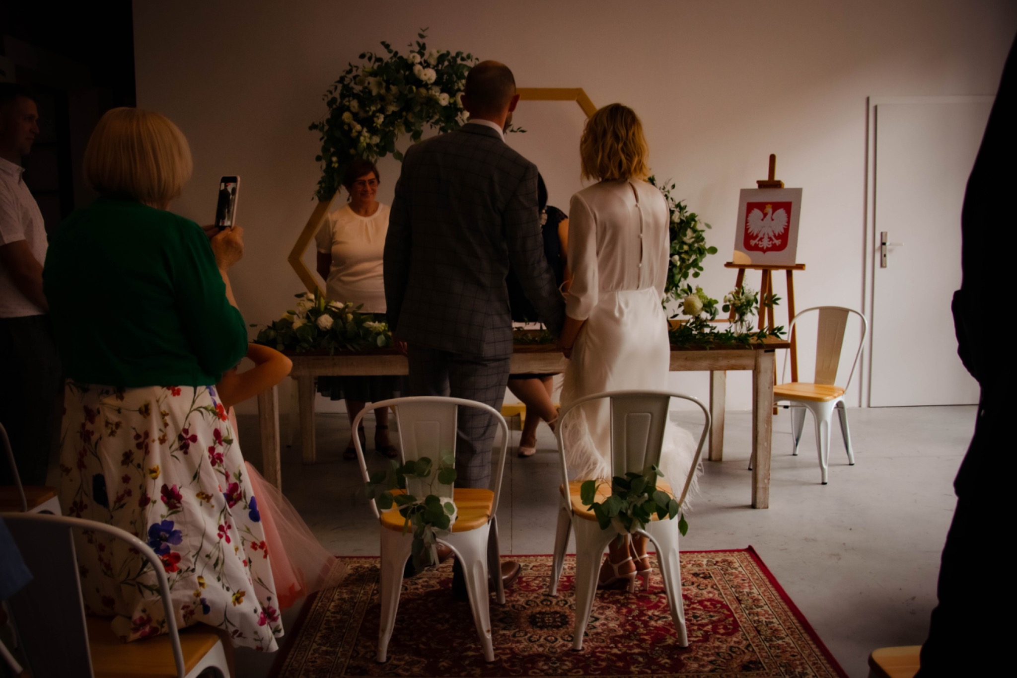 Para młoda stojąca przed heksagonalnym łukiem kwiatowym podczas ceremonii ślubnej, widok z tyłu, z flagą Polski na sztaludze w tle.