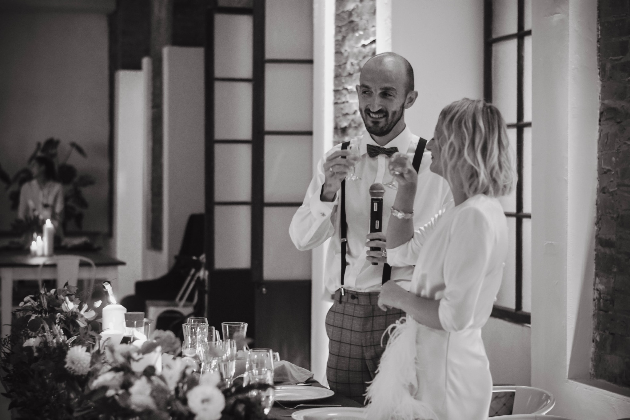 Czarno-białe zdjęcie mężczyzny w muszce i szelkach trzymającego mikrofon i toast, stojącego obok kobiety podczas przemówienia na tle stołu z kwiatami i świecami.