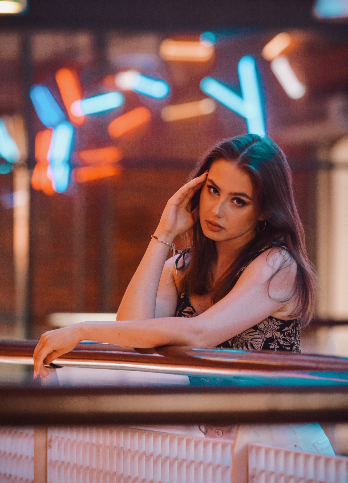 Portret kobiety opierającej się o metalową barierkę z neonowym oświetleniem w tle, bransoletka na nadgarstku, subtelny makijaż.