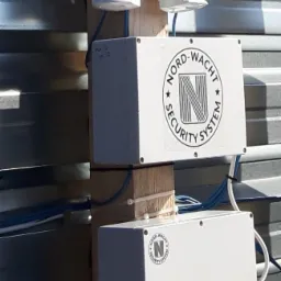 Dwa czujniki ruchu i dwie białe skrzynki z logo Nord-Wacht Security System zamontowane na drewnianej belce przy metalowym płocie.