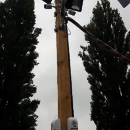 Drewniany słup z zamontowanymi reflektorami, kamerami i czujnikami ruchu, otoczony drzewami, na tle pochmurnego nieba.