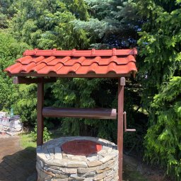 Artem Rozhko - Studnia z kamienia z drewnianą konstrukcją i czerwoną dachówką na tle zielonych drzew iglastych, widoczny fragment kostki brukowej i narzędzia budowlane w tle.