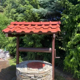 Studnia z kamienia z drewnianą konstrukcją i czerwoną dachówką na tle zielonych drzew iglastych, widoczny fragment kostki brukowej i narzędzia budowlane w tle.