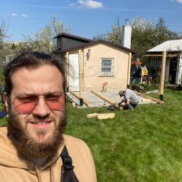Artem Rozhko - Mężczyzna w okularach i bluzie robi selfie na tle budowy małego domku z drewnianą konstrukcją pod podłogę i drugim pracownikiem w tle