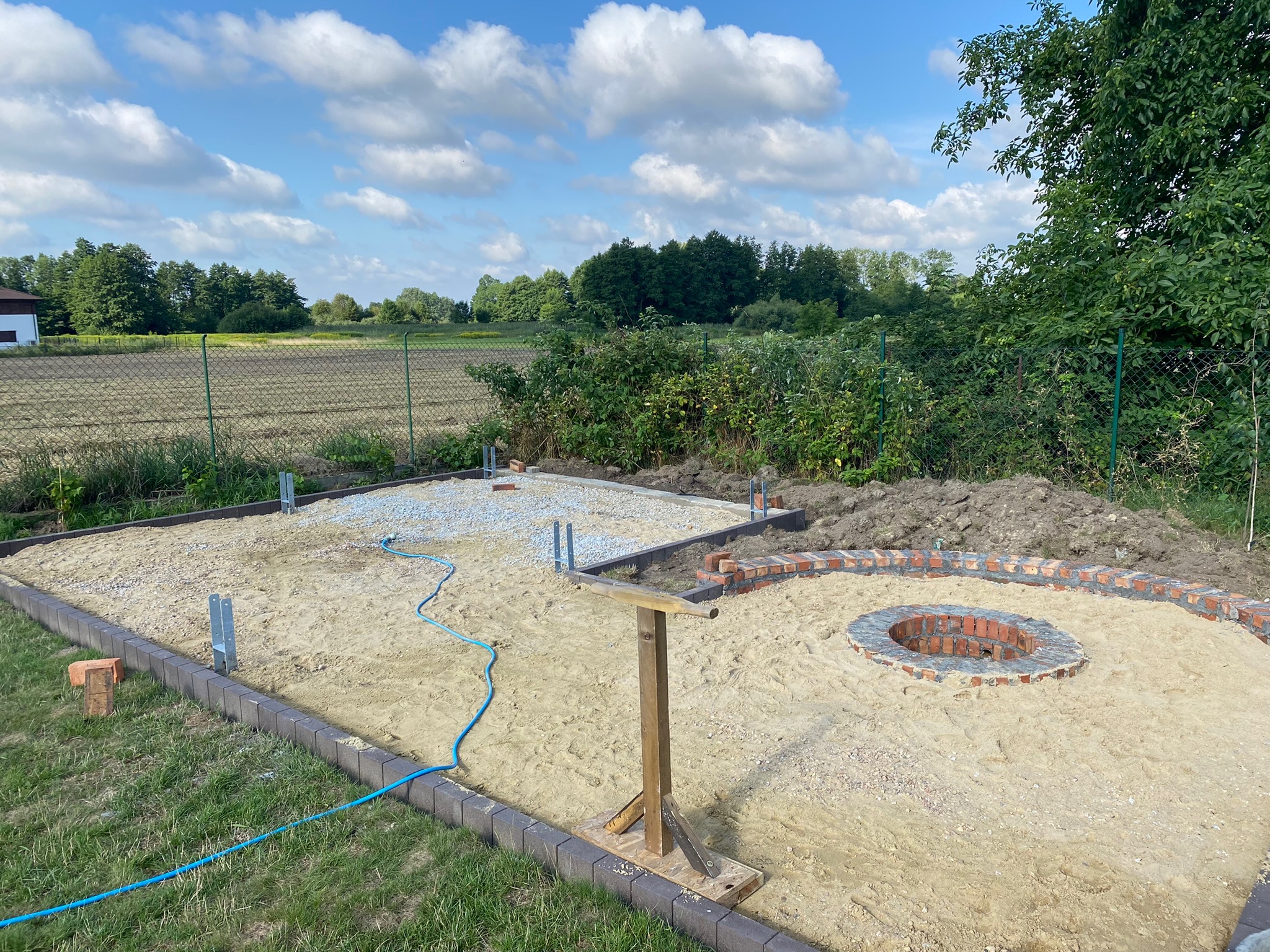 Przygotowanie podłoża pod budowę ogniska z cegły i tarasu z kostki, widoczne metalowe wsporniki, piasek, żwir i niebieski wąż ogrodowy.