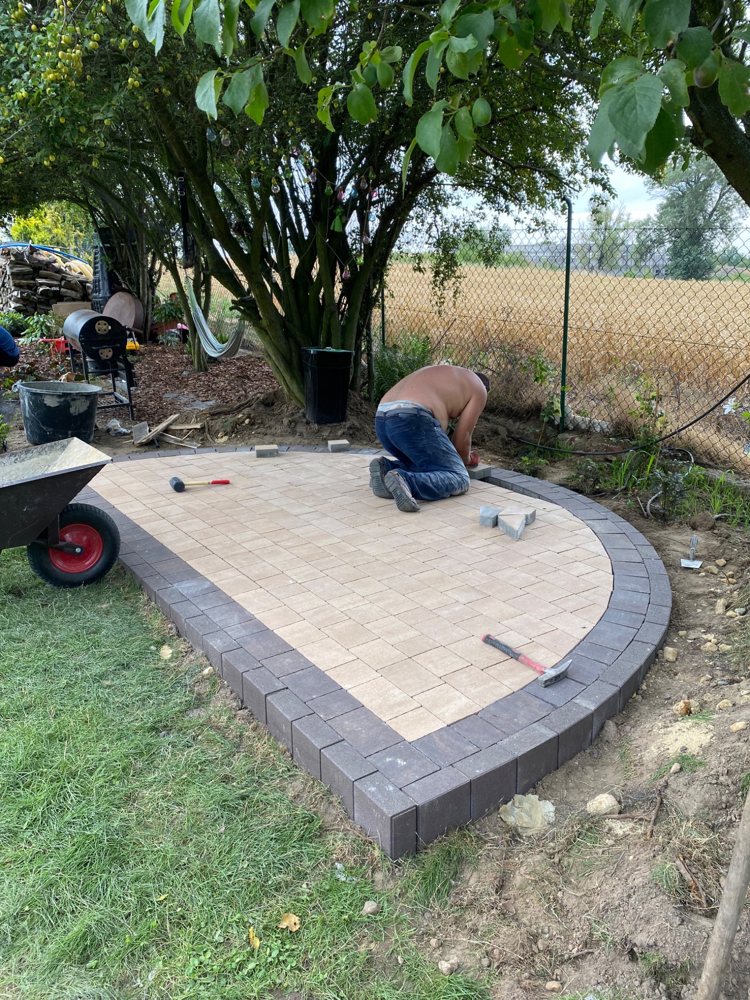 Układanie kostki brukowej na tarasie w ogrodzie, widoczny pracownik na kolanach, fragment taczki, teren częściowo zagospodarowany, w tle drzewa i siatka ogrodzeniowa.