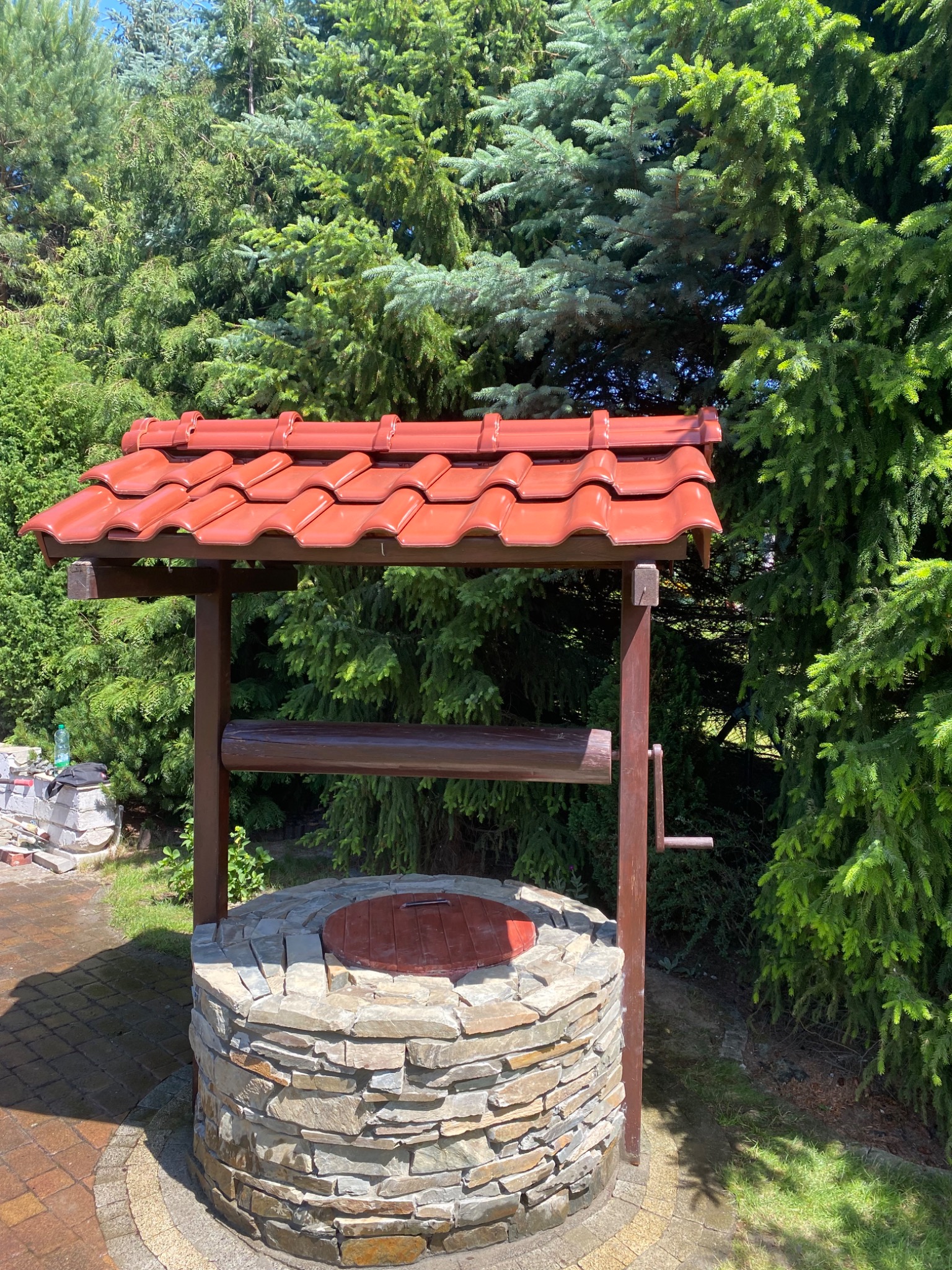Studnia z kamienia z drewnianą konstrukcją i czerwoną dachówką na tle zielonych drzew iglastych, widoczny fragment kostki brukowej i narzędzia budowlane w tle.