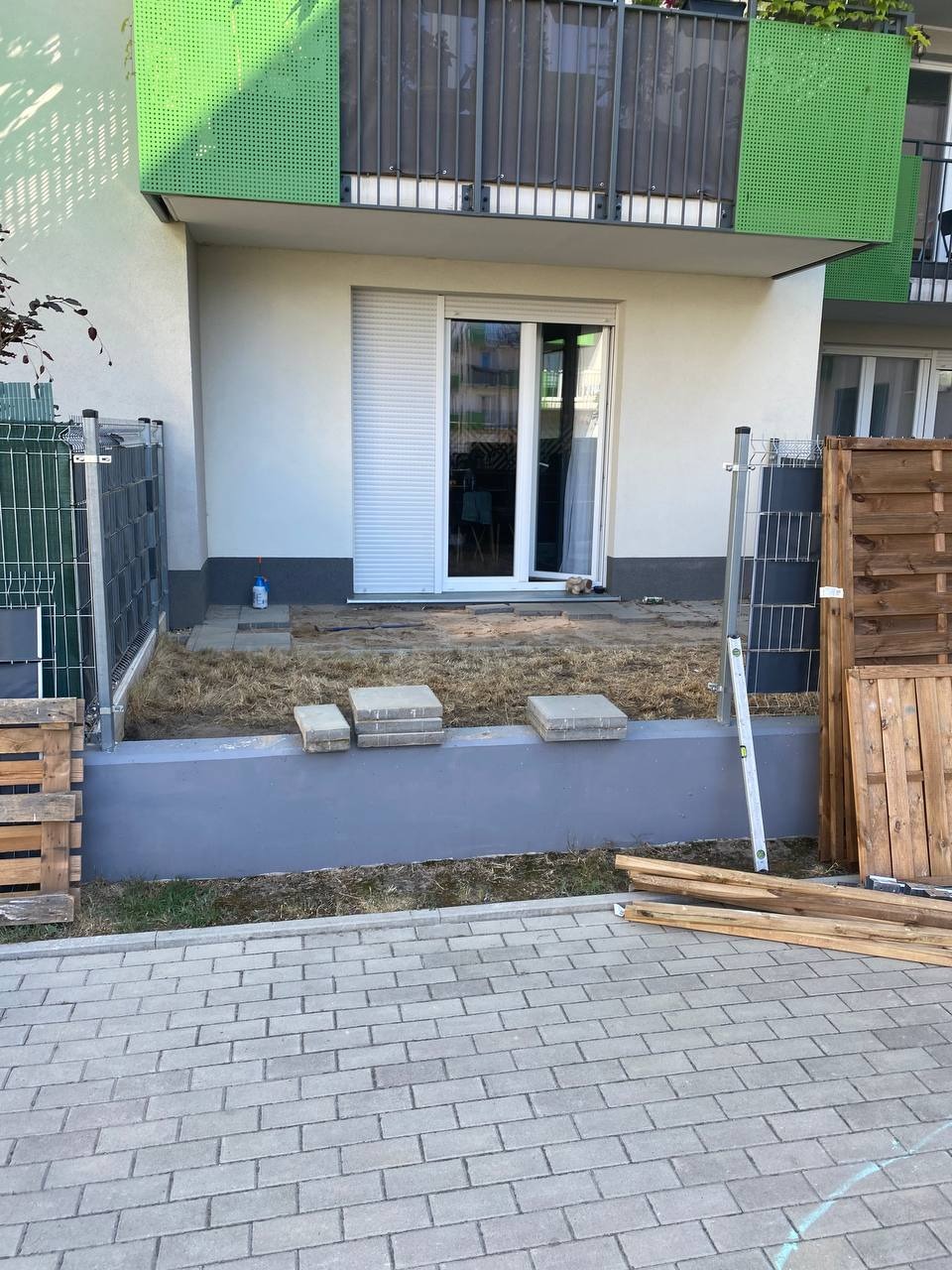 Przygotowanie do ułożenia kostki brukowej na tarasie przed budynkiem, widoczne palety, poziomica i ułożone płyty chodnikowe.