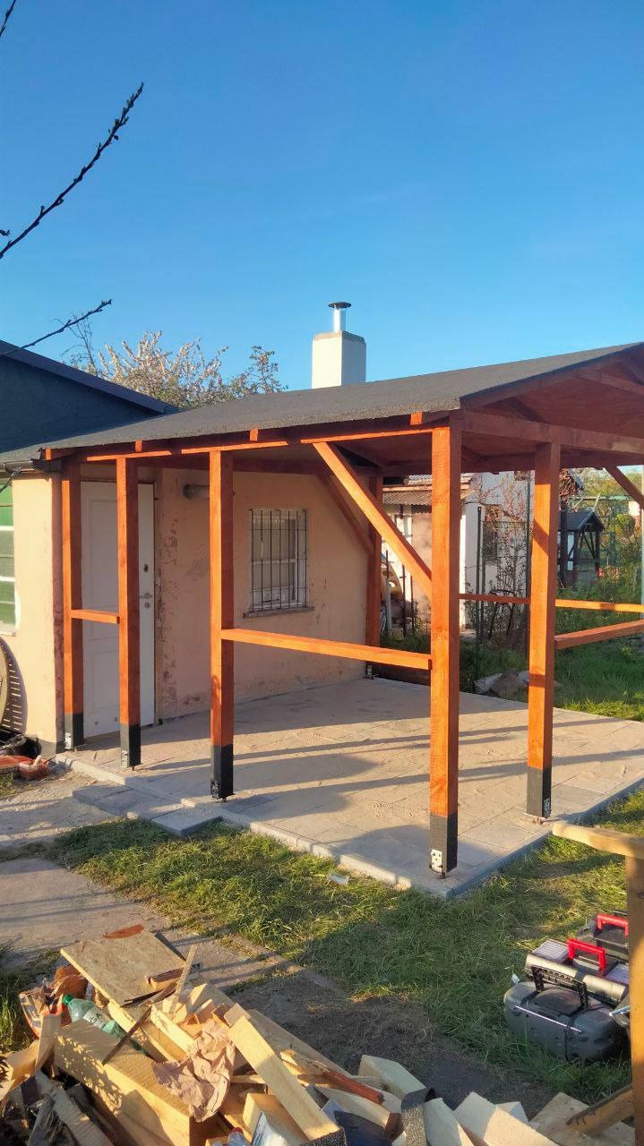 Drewniana konstrukcja z zadaszeniem w trakcie budowy, widoczne słupy z czarnymi podstawami, niekompletne barierki, na pierwszym planie stos drewna i narzędzia.