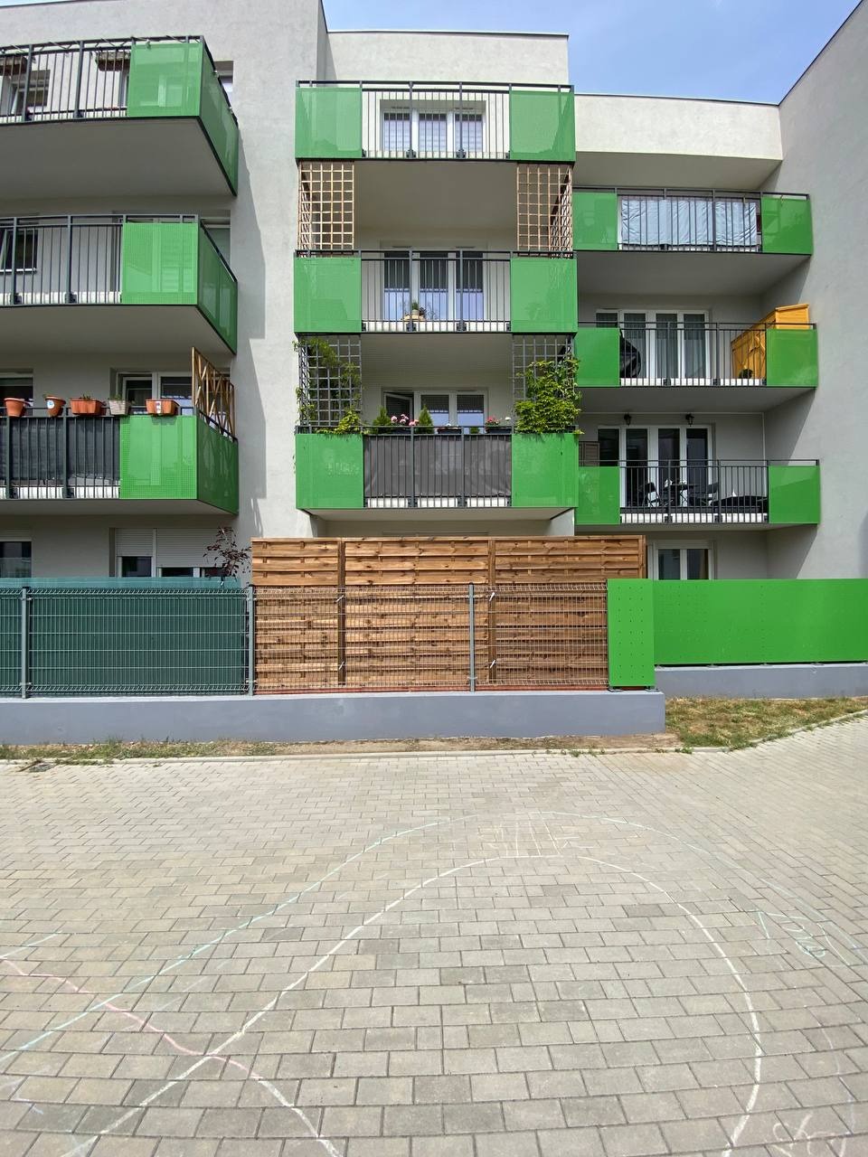 Nowoczesny budynek wielorodzinny z balkonami zabezpieczonymi zielonymi panelami, drewnianym ogrodzeniem i metalową siatką, widok z poziomu kostki brukowej z rysunkami kredą.