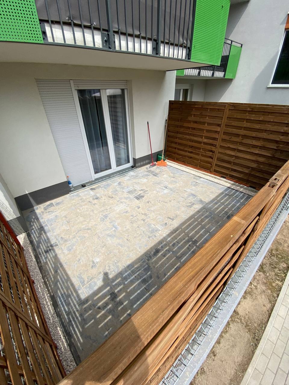 Nowo wyłożony, jasny taras z szarej kostki brukowej, obramowany drewnianym płotem i panelami, widok z góry, z widocznym cieniem konstrukcji.