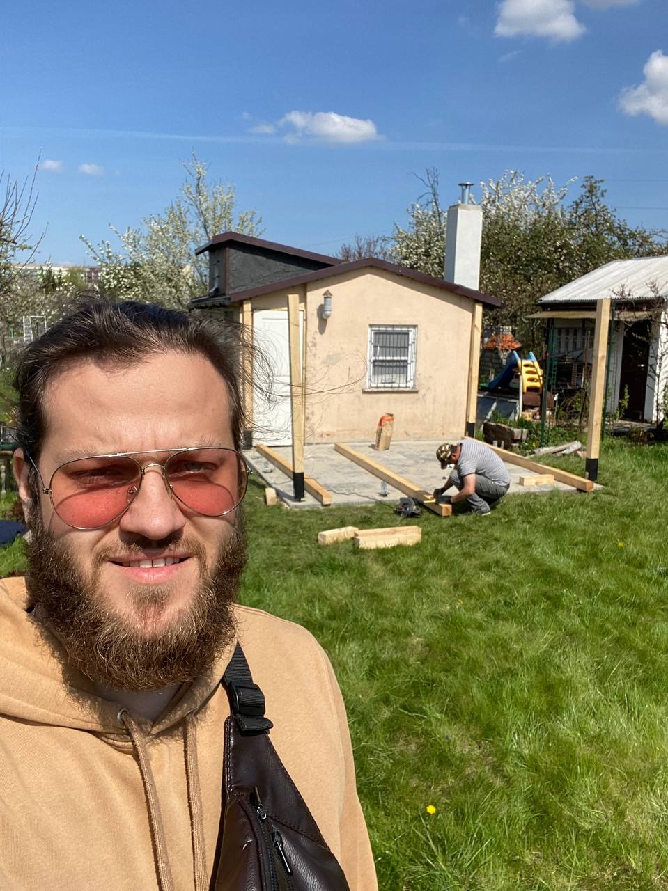 Mężczyzna w okularach i bluzie robi selfie na tle budowy małego domku z drewnianą konstrukcją pod podłogę i drugim pracownikiem w tle