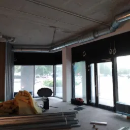 Wnętrze budynku komercyjnego w trakcie montażu wentylacji: widoczne rury wentylacyjne pod sufitem, stos wełny mineralnej, profile metalowe i duże okna.