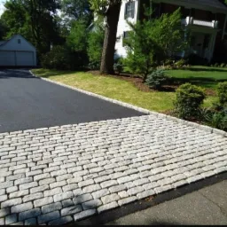 Podjazd do domu wyłożony jasnoszarą kostką brukową przechodzącą w asfaltową drogę, obrzeża z kostki, w tle zielony trawnik, drzewa i budynek mieszkalny.