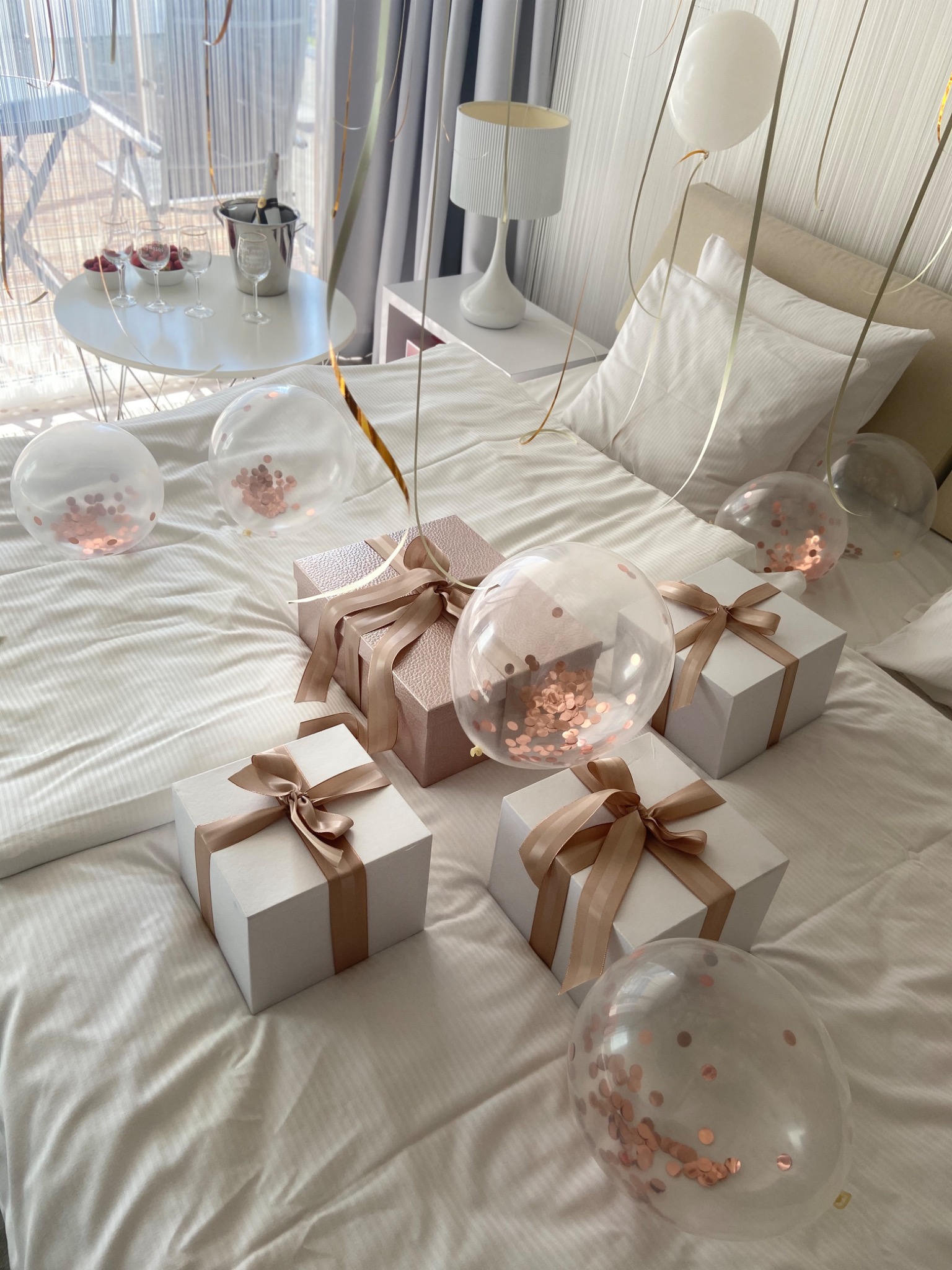 Prezenty w białych pudełkach z kokardami i przezroczyste balony z konfetti na białej pościeli, w tle stolik z szampanem i kieliszkami.