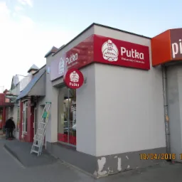 Zewnętrzny widok na budynek piekarni 'Putka' z dwoma czerwonymi szyldami z logo. Przy oknie oparta drabina aluminiowa, obok przechodnie. Widoczne inne lokale usługowe wzdłuż ulicy.