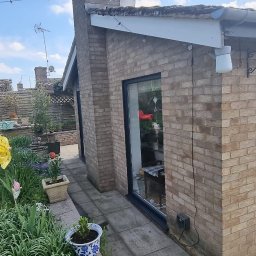 Homeefe Ltd - Widok z ogrodu na budynek z cegły z zamontowanymi przesuwnymi drzwiami w ciemnej ramie. W tle widoczna osoba.