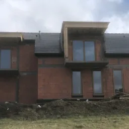 Surowa bryła domu w trakcie budowy z zamontowanymi oknami o ciemnych ramach, widoczny mur z czerwonej cegły, szara dachówka i ziemia na pierwszym planie.