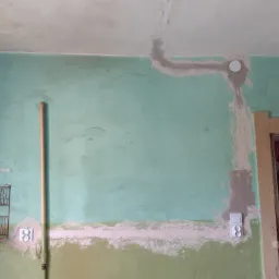 Ściana w trakcie remontu z nowymi gniazdkami elektrycznymi, widoczne ślady po kuciu i szpachlowaniu, rura pionowa i suszarka na naczynia po lewej stronie.