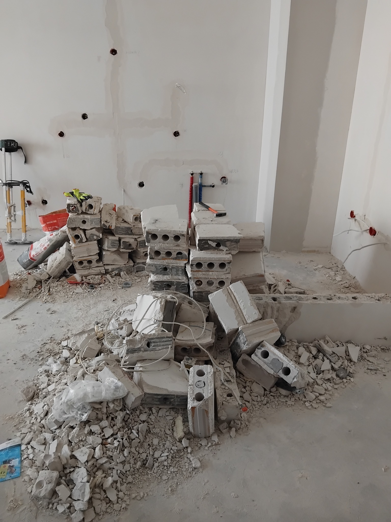 Stos białych pustaków i gruzu w surowym wnętrzu, widoczne instalacje elektryczne i hydrauliczne, narzędzia malarskie. Etap prac wykończeniowych w pomieszczeniu.