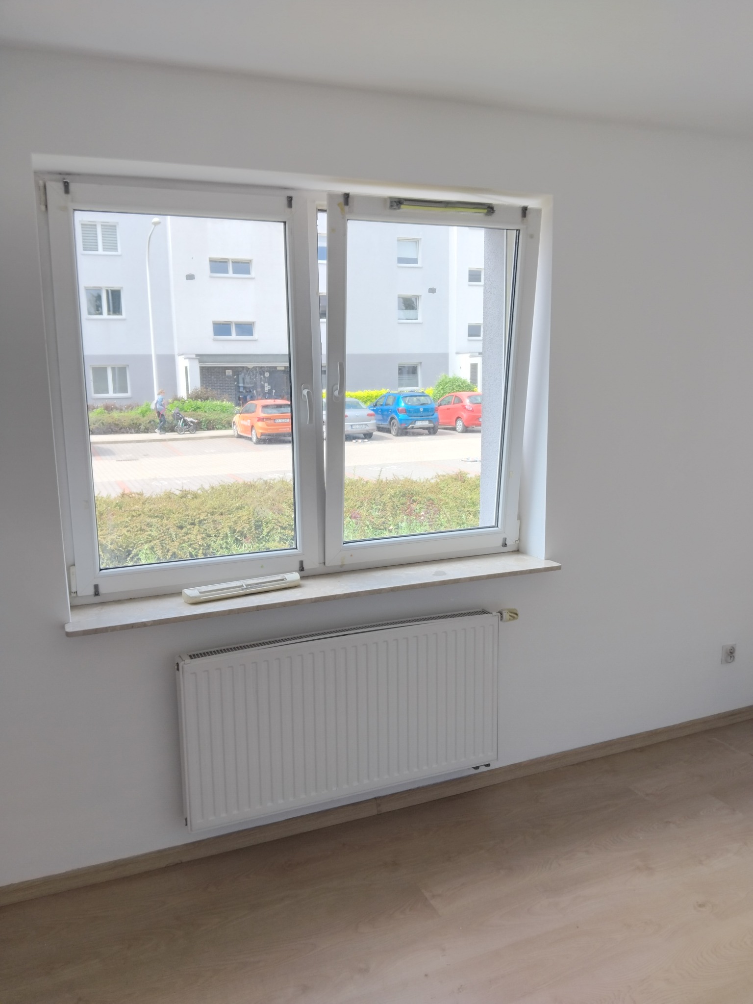 Białe ściany, okno z widokiem na parking z autami w tle, jasne panele podłogowe i grzejnik. Świeżo wykończone wnętrze mieszkania.
