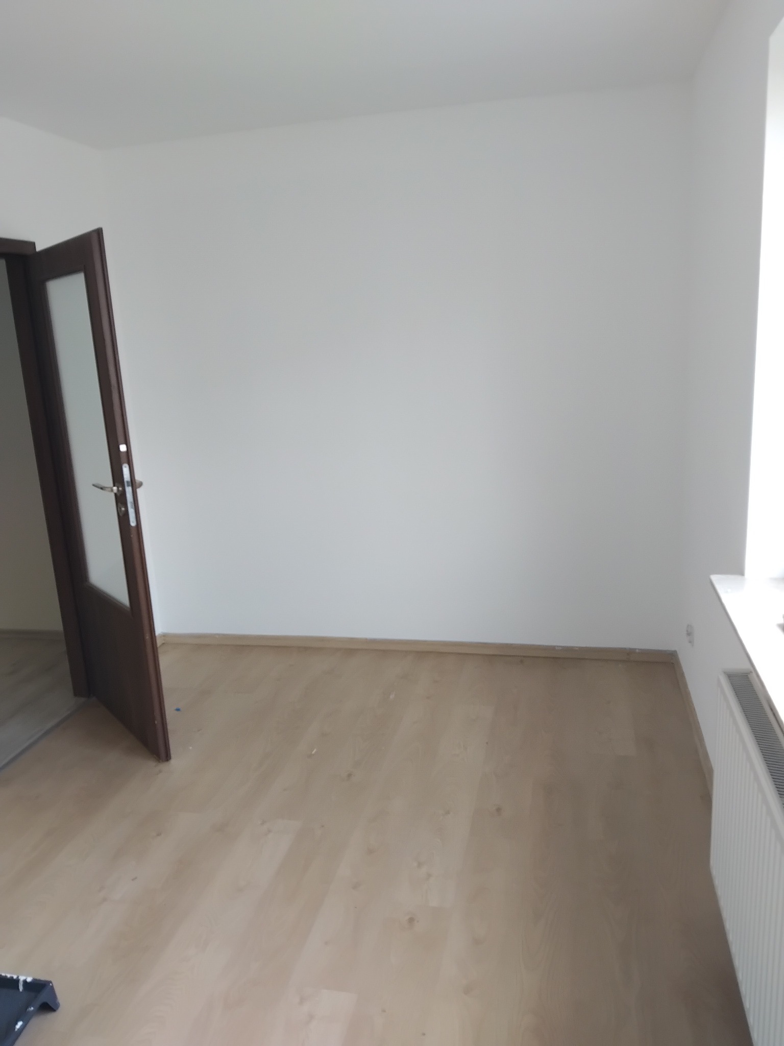 Jasne wnętrze po remoncie: świeżo pomalowane ściany, panele podłogowe i biały grzejnik przy oknie. Otwarta, ciemna stolarka drzwiowa z przeszkleniami.