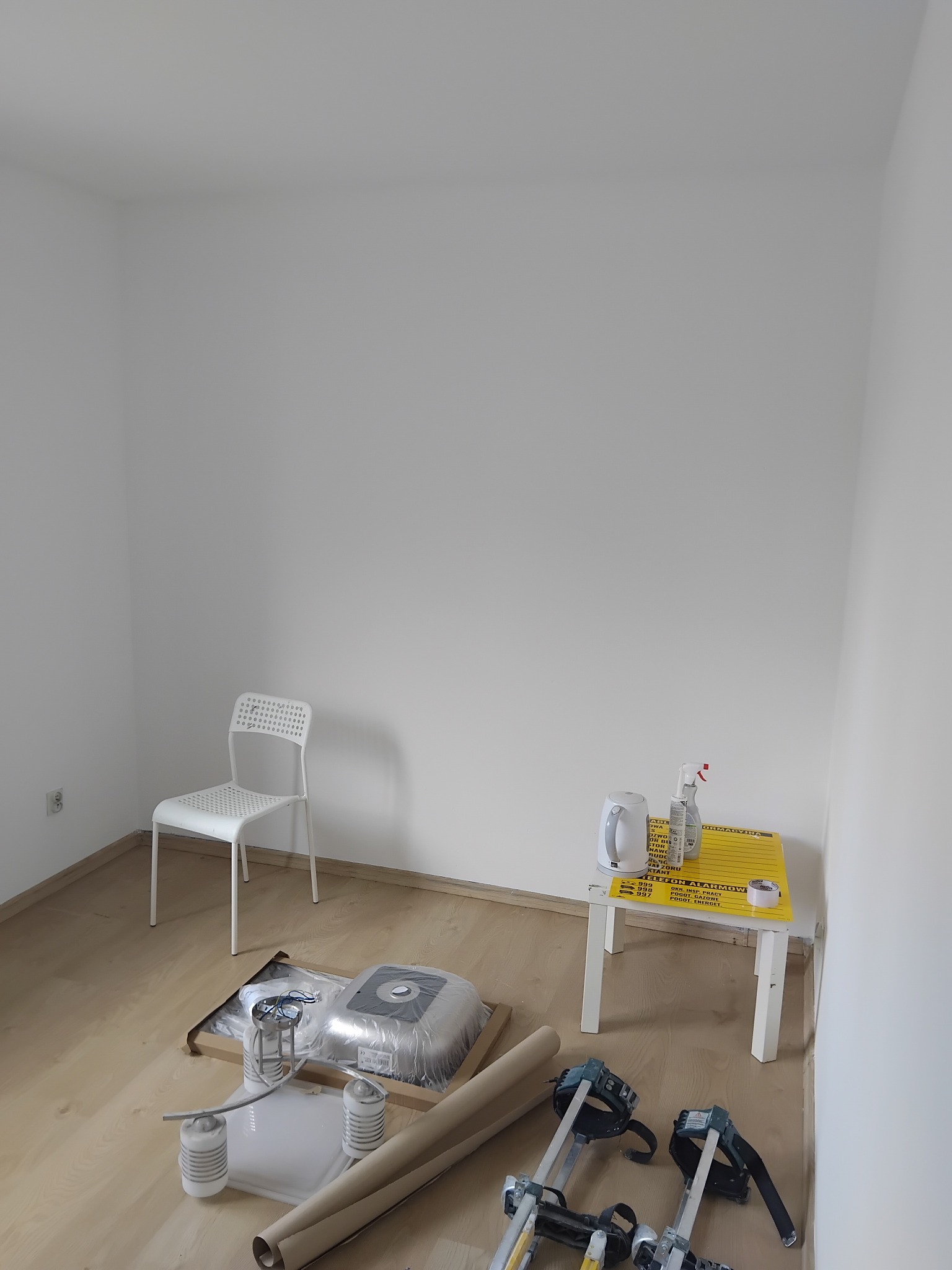 Przygotowania do remontu pokoju: białe ściany, krzesło, narzędzia malarskie, stół z czajnikiem i środkami czyszczącymi. Podłoga wyłożona panelami.