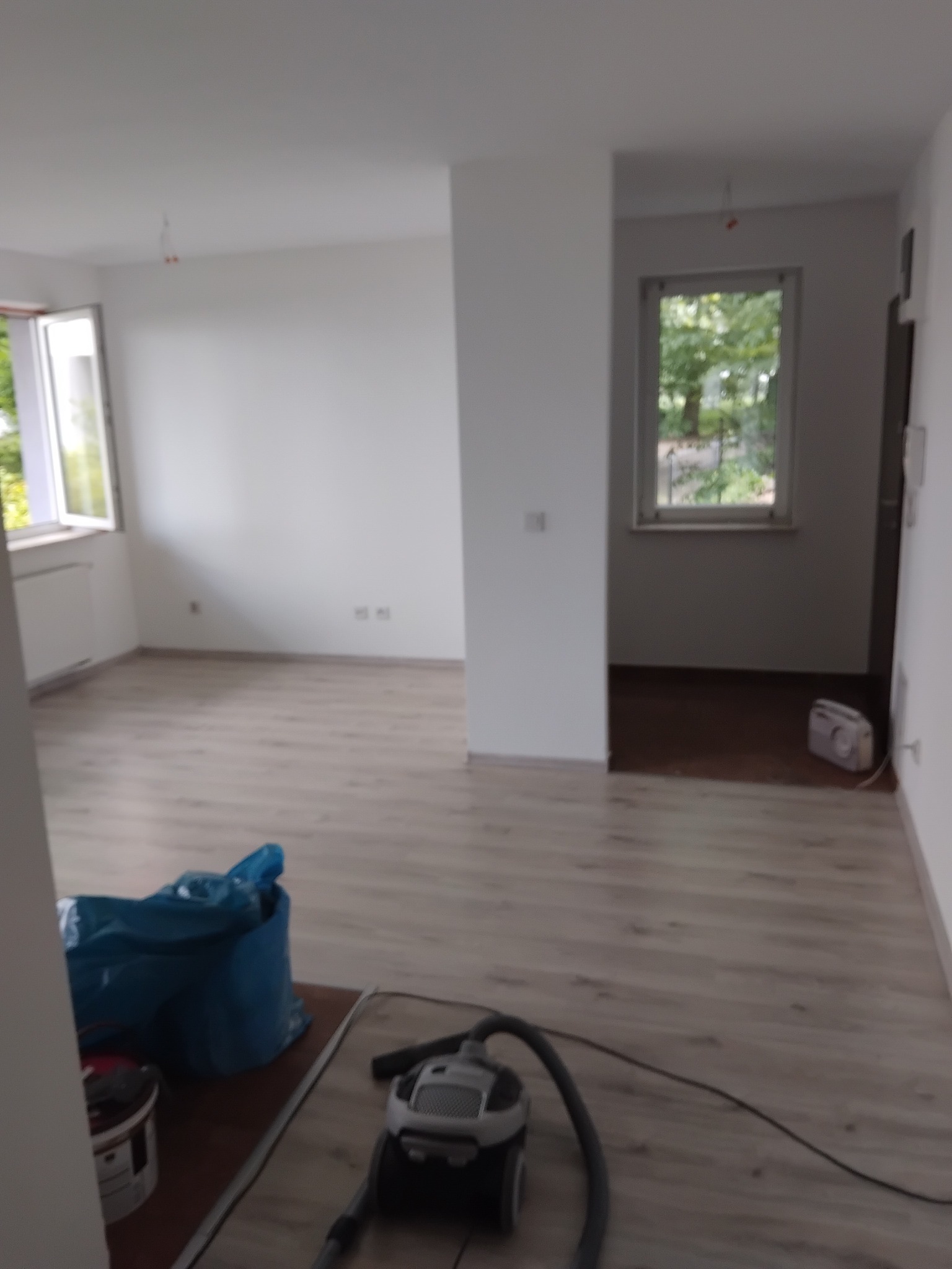 Jasne, odświeżone wnętrze po remoncie. Nowe panele podłogowe, świeżo pomalowane ściany, okno z widokiem na zieleń. Widoczny odkurzacz i worek z odpadami.