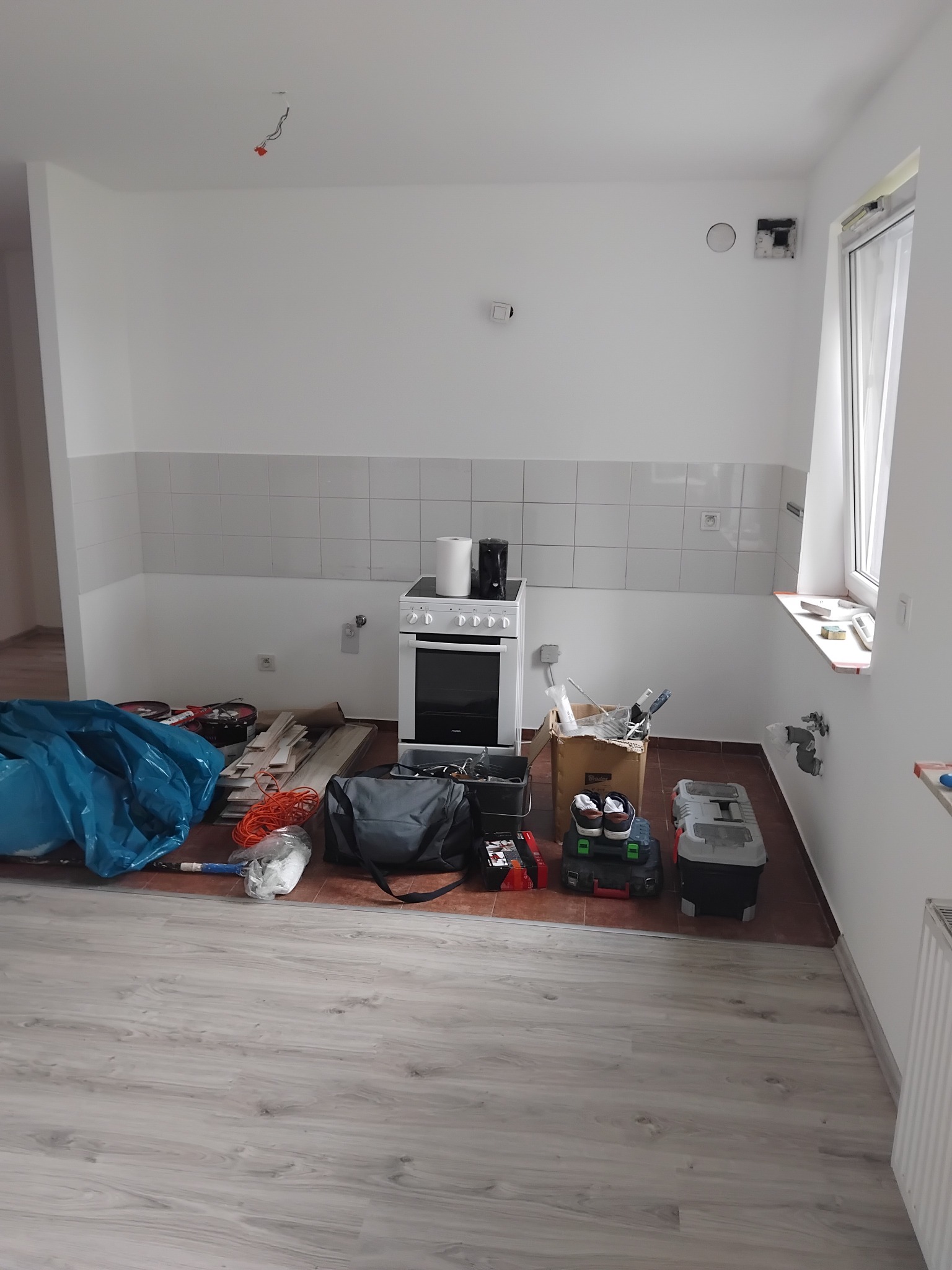 Wnętrze kuchni w trakcie prac wykończeniowych: białe ściany z płytkami, podłoga z paneli, narzędzia i materiały budowlane, kuchenka. Widoczne gniazdka elektryczne.