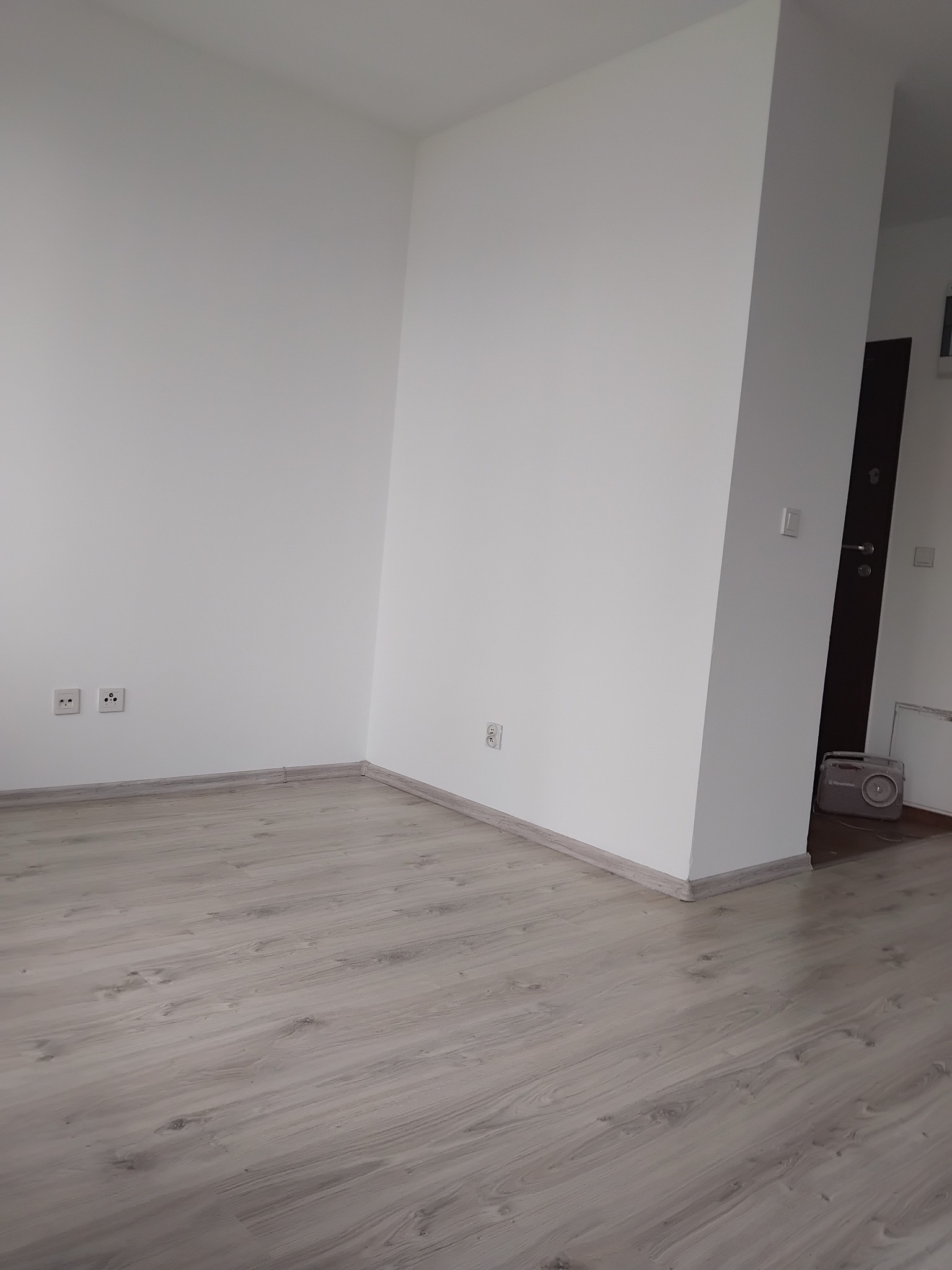 Jasne panele podłogowe w nowym, pomalowanym na biało pokoju. Widoczne gniazdka elektryczne i listwy przypodłogowe. W tle fragment drzwi i radia.