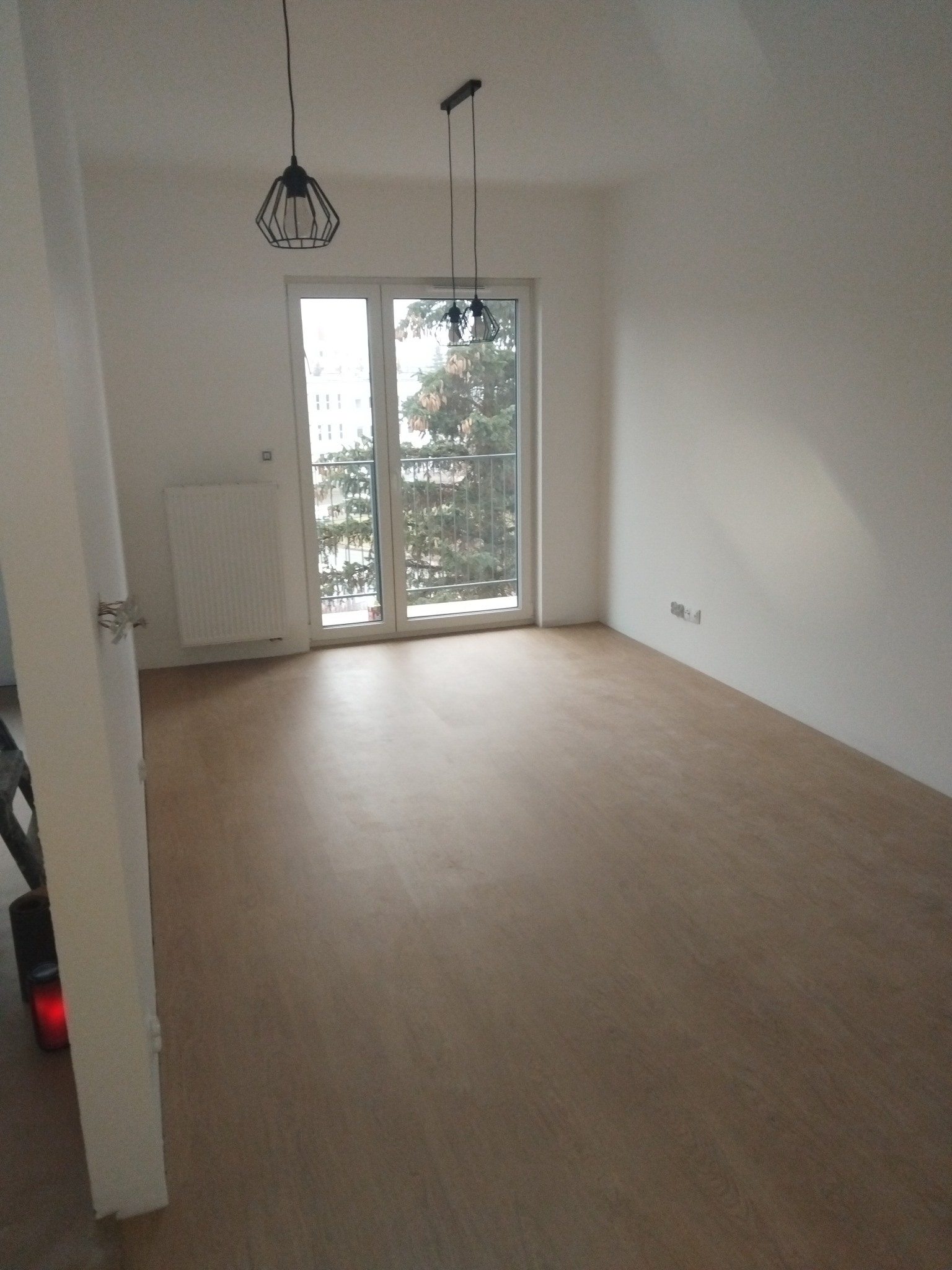 Jasne, puste wnętrze po remoncie: parkiet, białe ściany, okno balkonowe z widokiem na drzewa. Dwie wiszące lampy o geometrycznym designie.