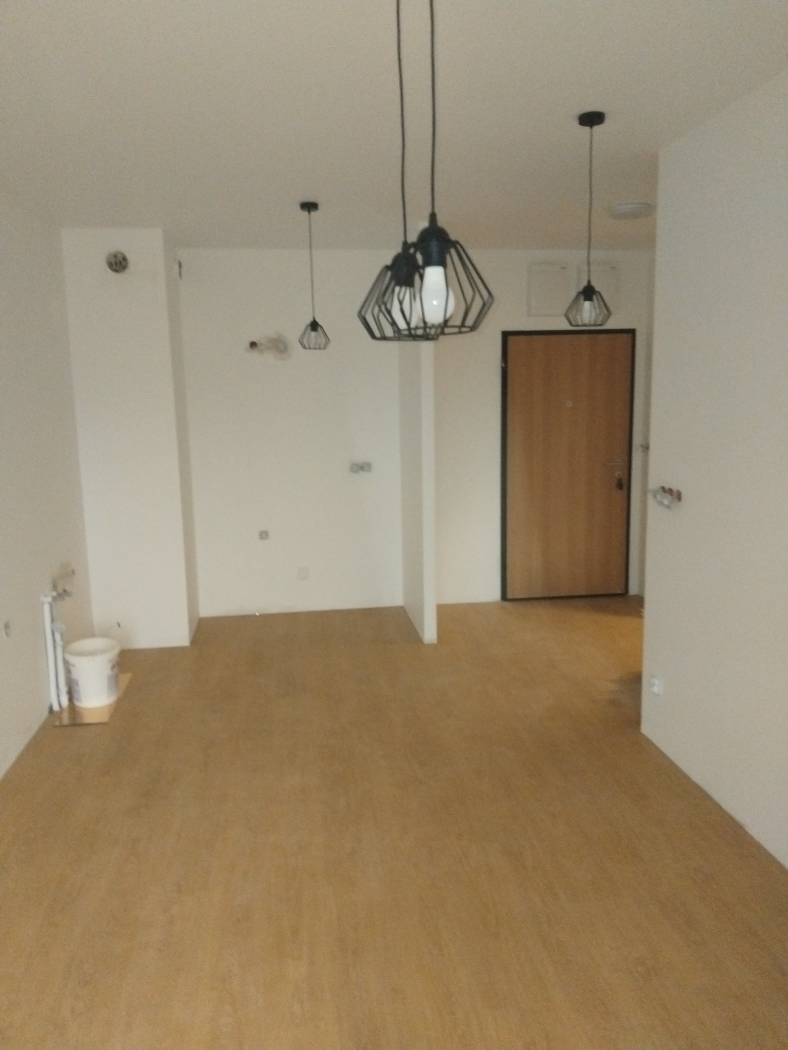 Jasne wnętrze po remoncie: panele podłogowe, pomalowane ściany na biało, wiszące lampy. Widoczne ślady po pracach wykończeniowych, np. wiadro z farbą.