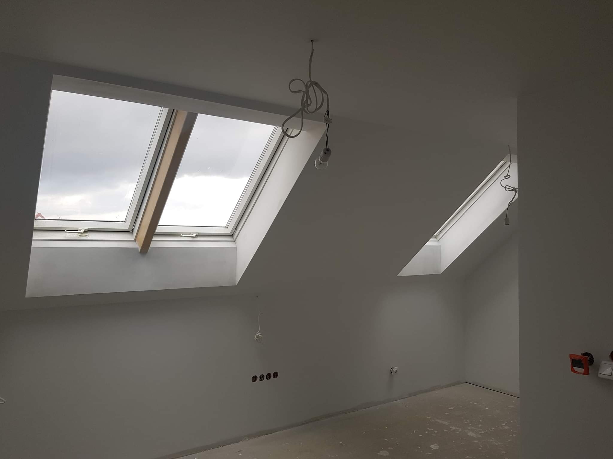 Poddasze po odświeżeniu ścian na biało, widoczne dwa okna dachowe z drewnianą ramą, surowy kabel z żarówką zwisający z sufitu, gniazdka elektryczne i przełączniki na ścianie.