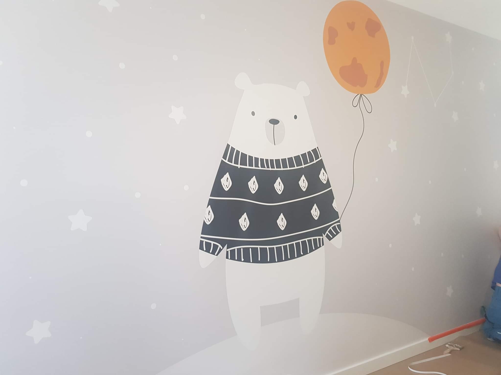 Ściana w pokoju dziecięcym z ręcznie malowanym motywem białego misia w swetrze trzymającego pomarańczowy balon, gwiazdki w tle, widoczny fragment listwy przypodłogowej i narzędzia malarskie.