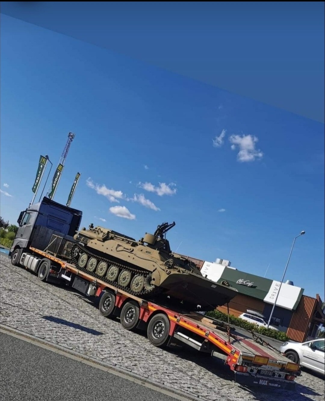 Ciężarówka z naczepą przewożąca transporter opancerzony na tle błękitnego nieba i budynku McDonald's Drive.