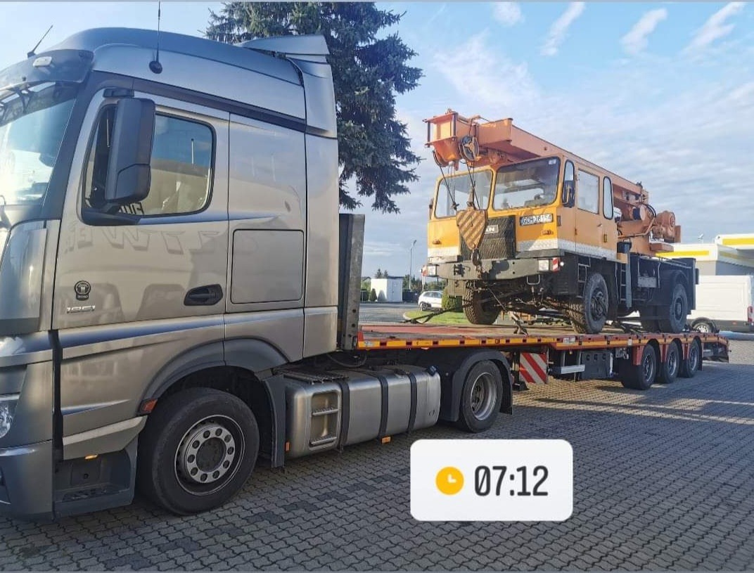 Ciężarówka z naczepą niskopodwoziową przewożąca dźwig samochodowy w kolorze pomarańczowym, widok z boku, na parkingu.