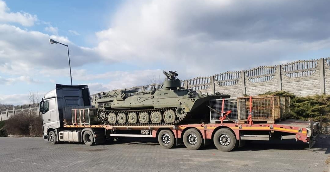 Ciężarówka z naczepą niskopodwoziową transportuje opancerzony transporter gąsienicowy na tle betonowego ogrodzenia i pochmurnego nieba.