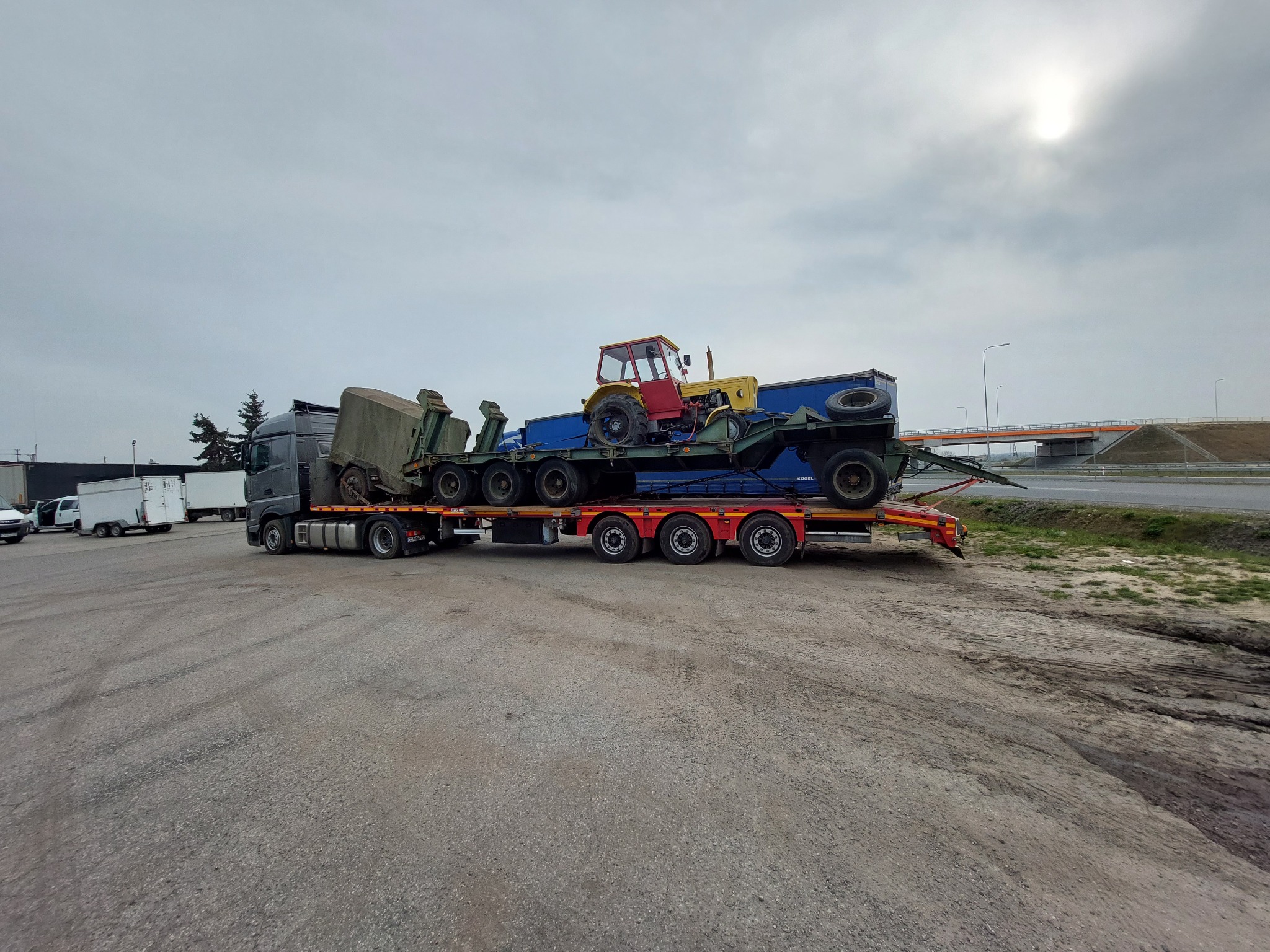 Ciężarówka z naczepą niskopodwoziową przewożąca traktor i elementy konstrukcyjne, widok z boku na placu manewrowym.