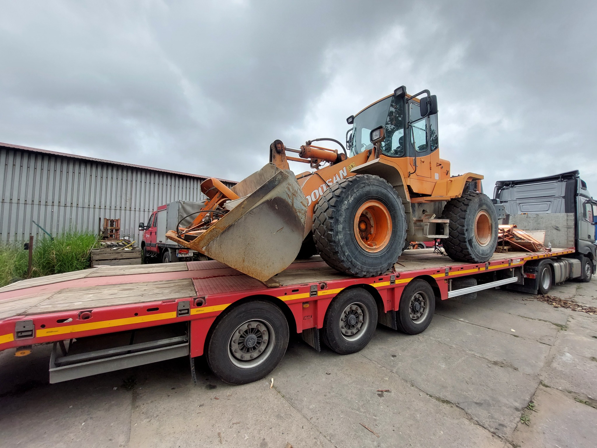 Pomarańczowa ładowarka kołowa Doosan przewożona na niskopodwoziowej platformie transportowej z czerwono-żółtymi elementami, widoczna z boku, na tle szarego nieba i budynków gospodarczych.