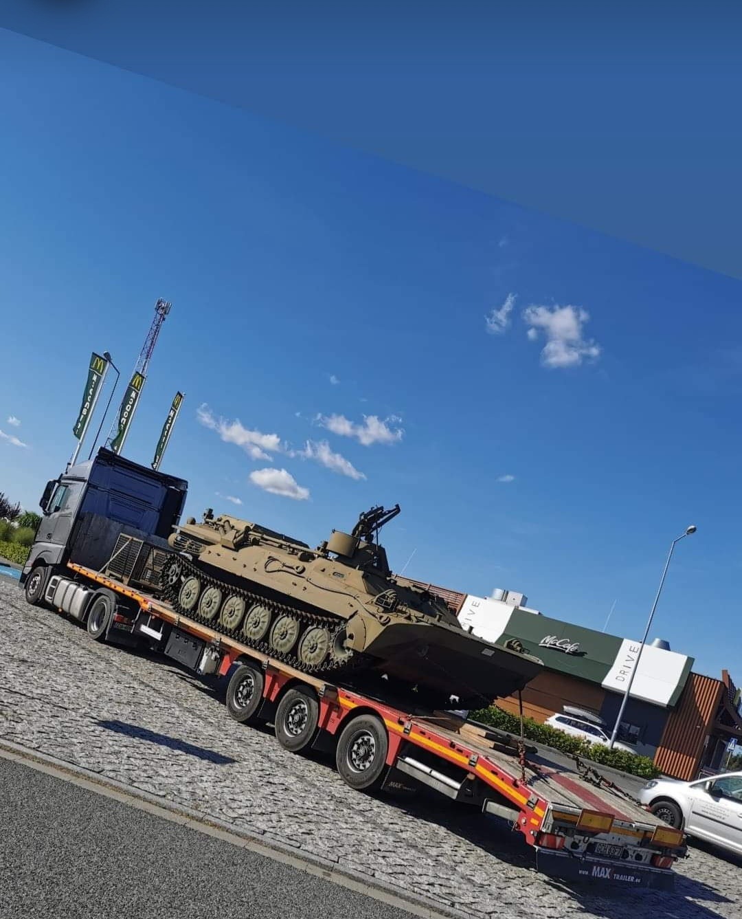 Ciężarówka z naczepą niskopodwoziową przewożąca pojazd wojskowy (BMP) na tle restauracji McDonald's.